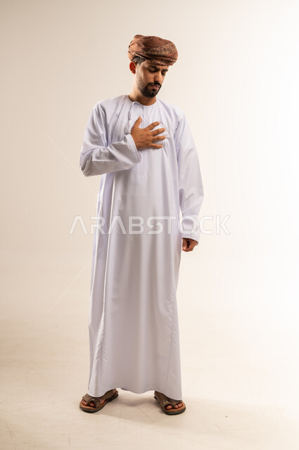 Feeling an increase in heart rate, the need to go to the doctor to conduct the necessary tests, heart disorders and diseases, a portrait of an Arab Gulf Omani man wearing a dishdasha and turban, placing his hand on his chest, feeling a tingling and pain in his heart, a full-length body image, white background