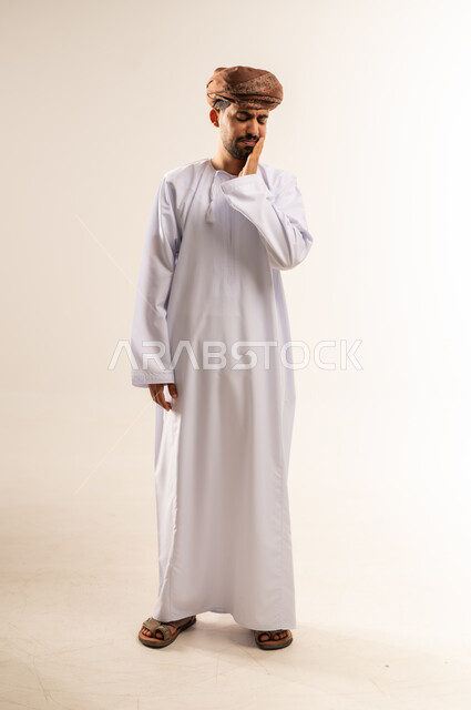 تسوس الأضراس وإلتهاب اللثه، طلب العلاج و المساعده، بورتريه لشاب عربي خليجي عماني يرتدي الدشداشة و العمامه يضع يده على خده ويغمض عينيه بإيماءات الشعور بألم الأسنان، الحاجه لزيارة الطبيب، صورة الجسم بالطول الكامل, خلفية بيضاء