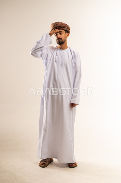 الاحساس بالشقيقية والصداع النصفي, الشعور بالإرهاق وفقدان التركيز, الاجهاد والضغط النفسي, بورتريه لشاب عربي خليجي عماني يرتدي الدشداشة و العمامه يضع يديه على رأسه ويغمض عينيه بإيماءات ألم الرأس الشديد, صورة الجسم بالطول الكامل, خلفية بيضاء