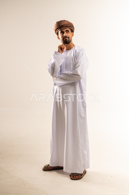 Deep thinking and absent-mindedness, trying to find a solution to difficult problems, anxiety and confusion in making the right decision, portrait of an Arab Gulf Omani man wearing a dishdasha and turban, putting his hand under his chin and looking somewhere far away with gestures of integration, full-length body, white background