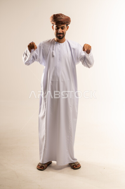 السرور والدهشة بوجود شيء ما، بورتريه لشاب عربي خليجي عماني مبتسم يرتدي الدشداشة و العمامة يشير بأصابعه نحو الأسفل بإيماءات تدل على هذا المكان (هنا)، صورة الجسم بالطول الكامل، خلفية بيضاء