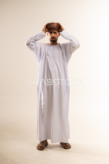 آلام الصداع النصفي، التعرض لنوبات الشقيقة الحادة، الحاجة لتناول المسكنات وأخذ قسط من الراحة، التعرض للتعب والإرهاق والعمل الطويل، بورتريه لرجل عربي خليجي عماني مبتسم يرتدي الدشداشة والعمامة يمسك رأسه بيديه بإيماءات الألم، الجسم بالطول الكامل، خلفية بيضاء