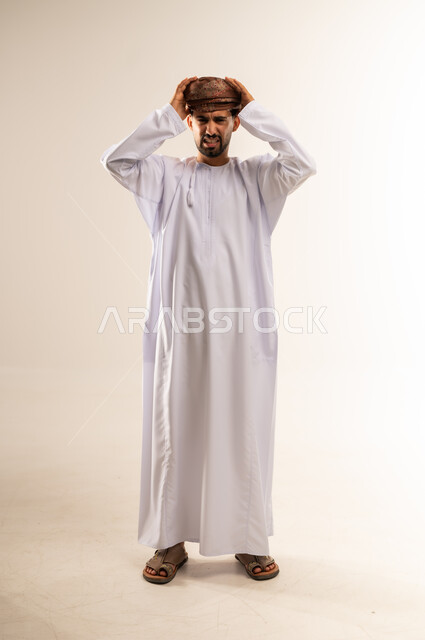 آلام الصداع النصفي، التعرض لنوبات الشقيقة الحادة، الحاجة لتناول المسكنات وأخذ قسط من الراحة، التعرض للتعب والإرهاق والعمل الطويل، بورتريه لرجل عربي خليجي عماني مبتسم يرتدي الدشداشة والعمامة يمسك رأسه بيديه بإيماءات الألم، الجسم بالطول الكامل، خلفية بيضاء