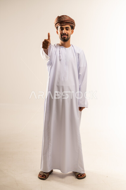 تعابير الموافقة والرضى والقبول، بورتريه لشاب عربي خليجي عماني مبتسم يرتدي الدشداشة و العمامة يقف و يرفع اصبع الابهام للاعلى بإيماءات الاعجاب والسرور، صورة الجسم بالطول الكامل، خلفية بيضاء