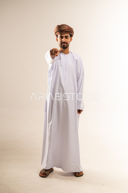 اشارات تحدد هذا المكان (هنا)، الاشارة إلى شيء ما، تعابير التحديد والتأكيد، بورتريه لرجل عربي خليجي عماني مبتسم يرتدي الدشداشه والعمامة يشير بإصبع السبابة وينظر الى الكاميرا بإيماءات السعادة والسرور، الجسم الكامل بالطول، خلفية بيضاء