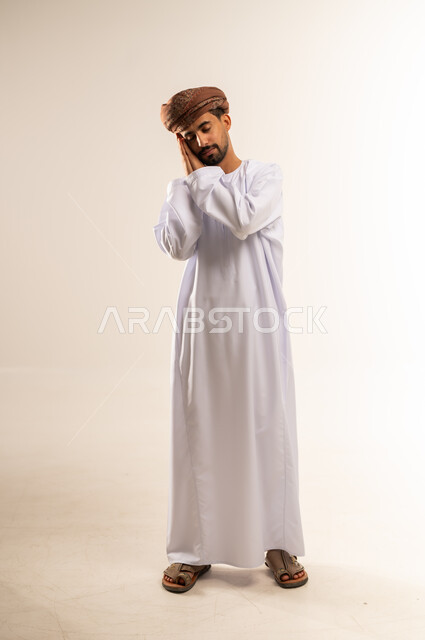 الشعور بالتعب و الحاجه الى الراحه, الإحساس بالكسل والخمول، بورتريه  لشاب عربي خليجي عماني يرتدي الدشداشة و العمامة يقف ويضع يديه تحت رأسه ويغمض عينيه بإيماءات النوم، تعابير النعاس والإرهاق، الجسم الكامل بالطول، خلفية بيضاء