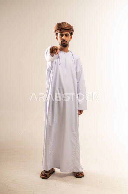 الإشارة باصبع السبابة إلى الكاميرا بشكل مباشر، التهديد والتوعد بتنفيذ شيء ما، إيماءات الترهيب والتحذير، توجيه أصابع الاتهام إلى شخص معين، بورتريه لرجل عربي خليجي عماني يرتدي الدشداشه والعمامة يشير إلى مكان ما محدد، الجسم بالطول الكامل، خلفية بيضاء