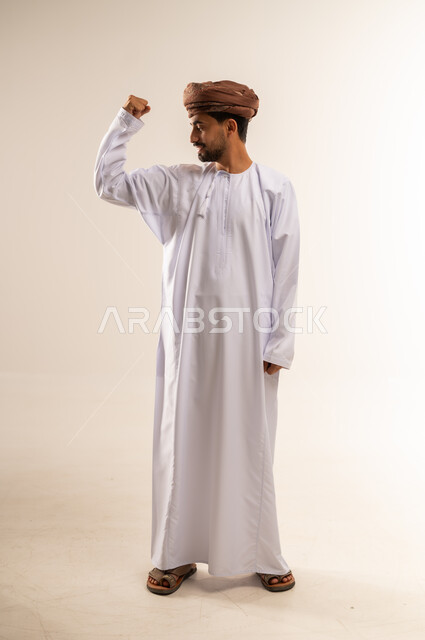 المواظبة على التمارين الرياضية، الاهتمام باللياقة الجسدية، اتباع نظام غذائي صحي، بناء الكتلة العضلية للجسم، مفهوم القوة والرجولة، بورتريه لرجل عربي خليجي عماني مبتسم يرتدي الدشداشه والعمامة ينظر لعضلاته بإيماءات السعادة، الجسم بالطول الكامل، خلفية بيضاء