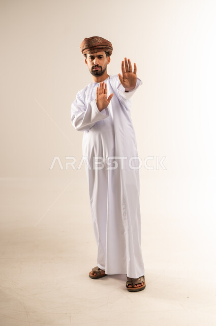 Request to stop doing something, order to stop and not go too far, refusal and not submit, prohibition of progress and importance of leaving a safe distance, portrait of an Arab Gulf Omani man wearing a dishdasha and turban raising his palm up with gestures of anger and seriousness, full body, white background