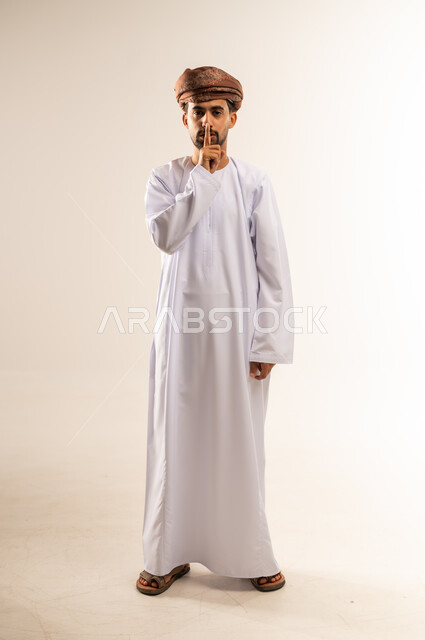 طلب التوقف عن الكلام والصمت، النهي والزجر والانصات، وضع اصبع السبابة أمام الفم بإيماءات الجدية، الأمر بالصمت التام، بورتريه لرجل عربي خليجي عماني يرتدي الدشداشه والعمامة ينظر للكاميرا بإيماءات الغضب، الجسم بالطول الكامل، خلفية بيضاء