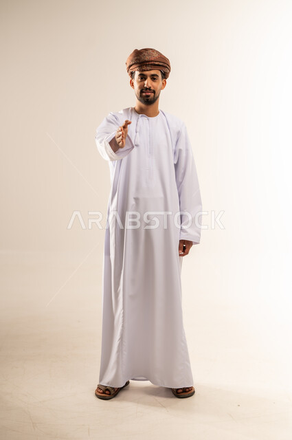 مد كف يد نحو الأمام للمصافحة، إلقاء التحية والسلام، الأناقة والوسامة والرجولة، مفهوم التقدير والاحترام، بورتريه لرجل عربي خليجي عماني مبتسم يرتدي الدشداشه والعمامة ينظر إلى الكاميرا بإيماءات السعادة والسرور، الجسم بالطول الكامل، خلفية بيضاء