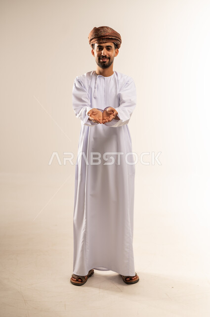 فتح الكفين ومدهما نحو الأمام، تقديم الضيافة أو إعطاء شيء ما، الأناقة والوسامة والرجولة، مفهوم التقدير والاحترام، بورتريه لرجل عربي خليجي عماني مبتسم يرتدي الدشداشه والعمامة ينظر إلى الكاميرا بإيماءات السعادة والسرور، الجسم بالطول الكامل، خلفية بيضاء