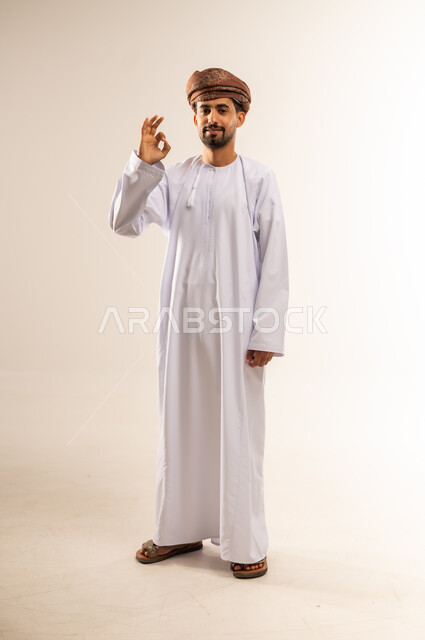 التعبير عن الإعجاب والتقدير، رفع اليد بعلامات الجودة والقبول، الامتياز والعلامة الكاملة، الشعور بالرضا والاستحسان، بورتريه لرجل عربي خليجي عماني مبتسم يرتدي الدشداشة والعمامة ينظر إلى الكاميرا بإيماءات السعادة والسرور، الجسم بالطول الكامل، خلفية بيضاء