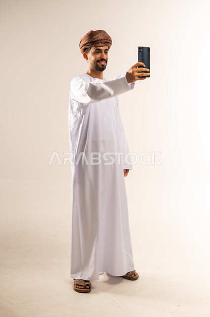 دمج التكنولوجيا والاجهزة الحديثة في الحياة اليومية، بورتريه لرجل عربي خليجي عماني مبتسم يرتدي الدشداشه والعمامة يقف ويقوم بإلتقاط صور سيلفي تذكارية باستخدام الهاتف المحمول، التواصل مع الاهل والاصدقاء، خلفية بيضاء