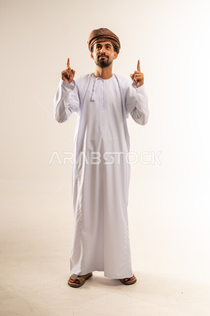 حركات يدين واشارات تدل على هذا المكان (هنا)، تحديد موقع شيء معين، بورتريه لرجل عربي خليجي عماني يرتدي الدشداشة والعمامة يشير باصبعيه السبابة إلى الأعلى، مفهوم الأناقة والاهتمام بالمظهر الخارجي، الجسم بالطول الكامل، خلفيه بيضاء