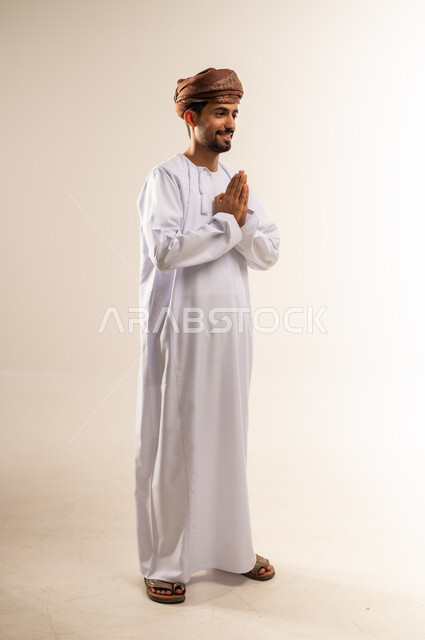 مفهوم الاستقبال والترحيب، الإكثار من الدعاء في شهر رمضان المبارك، الإجتهاد في العبادة والحفاظ على الطاعات، بورتريه لشاب عربي خليجي عماني مبتسم يرتدي الدشداشة والعمامة يرفع يديه ويدعو الله بإيماءات التوسل والتضرع، صورة الجسم بالطول الكامل, خلفية بيضاء