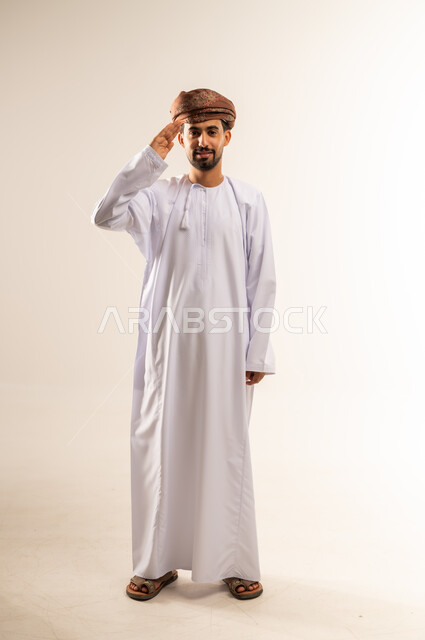 إلقاء التحية والسلام، الأناقة والوسامة والرجولة، مفهوم التقدير والاحترام، بورتريه لرجل عربي خليجي عماني مبتسم يرتدي الدشداشه والعمامة ينظر إلى الكاميرا بإيماءات السعادة والسرور، الوقوف باستقامة بتعابير الثقة بالنفس، الجسم بالطول الكامل، خلفية بيضاء