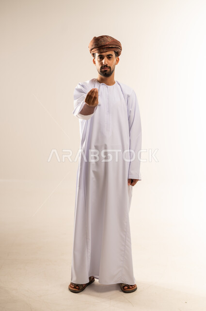 مفهوم الصبر والتريث و التأني, التهديد و التوعد لشيء ما، بورتريه لشاب عربي خليجي عماني يرتدي الدشداشة و العمامة ينظر الى الكاميرا ويرفع يده نحو الأعلى بإيماءات طلب الإنتظار, إمهال بعض الوقت لفعل شيء، صورة الجسم بالطول الكامل, خلفية بيضاء