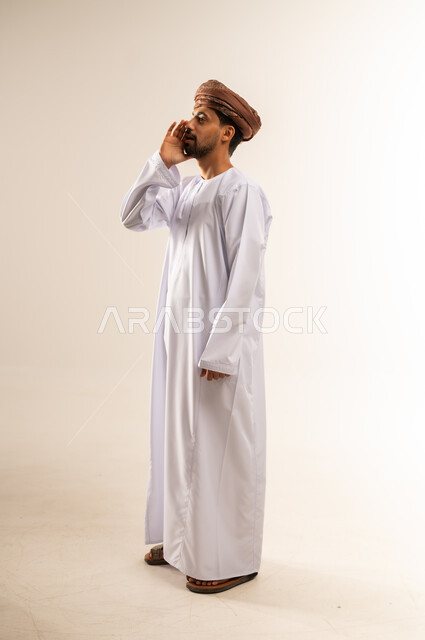 النداء والكلام والتحدث بصوت مرتفع، بورتريه من الجانب لشاب عربي خليجي عماني يرتدي الدشداشة و العمامة يضع يده على فمه بإيماءات المناداة لشخص ما، دردشة ومحادثات وتواصل اجتماعي، صورة الجسم بالطول الكامل, خلفية بيضاء