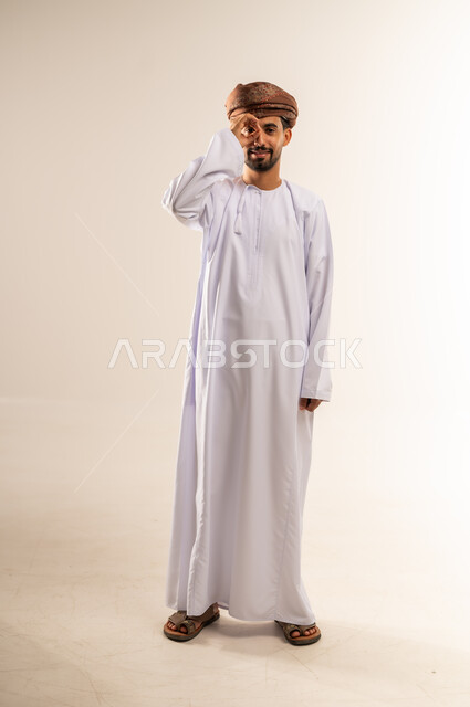 مفهوم التخطيط و الرؤية المستقبلية، بورتريه لشاب عربي خليجي عماني مبتسم يرتدي الدشداشة و العمامة يضع يده على عينيه بشكل عدسة دائرية بإيماءات البحث والاستكشاف، صورة الجسم بالطول الكامل, خلفية بيضاء