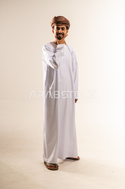 The concept of handsomeness and self-confidence, standing and looking at the camera with gestures of pleasure and cheerfulness, portrait of a smiling Arab Gulf Omani man wearing a dishdasha and turban, standing and placing his hand under his chin with a smiling expression, authentic Arab masculinity, interest in external appearance, full-length body, white background