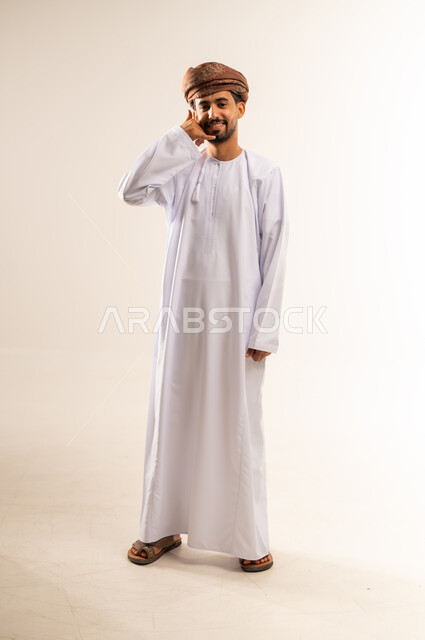 إنتظار مكالمة وإستخدام الميكروفون، بورتريه لشاب عربي خليجي عماني مبتسم يرتدي الدشداشة و العمامة يرفع يده بإيماءات الإتصال وإجراء مكالمة هاتفية، تعابير الفرح والسرور، صورة الجسم بالطول الكامل, خلفية بيضاء