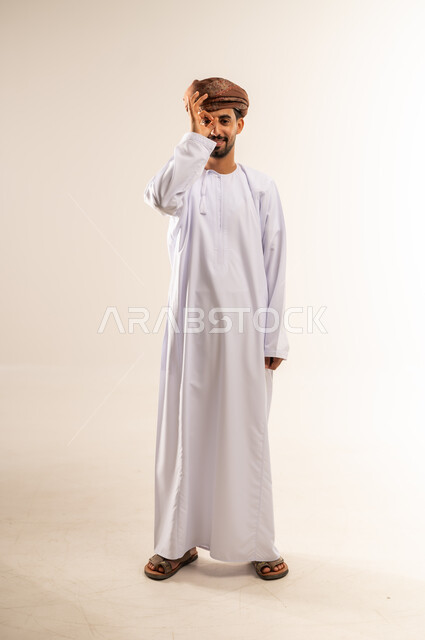 مفهوم التخطيط و الرؤية المستقبلية، بورتريه لشاب عربي خليجي عماني مبتسم يرتدي الدشداشة و العمامة يضع يده على عينيه بشكل عدسة دائرية بإيماءات البحث والاستكشاف، صورة الجسم بالطول الكامل, خلفية بيضاء