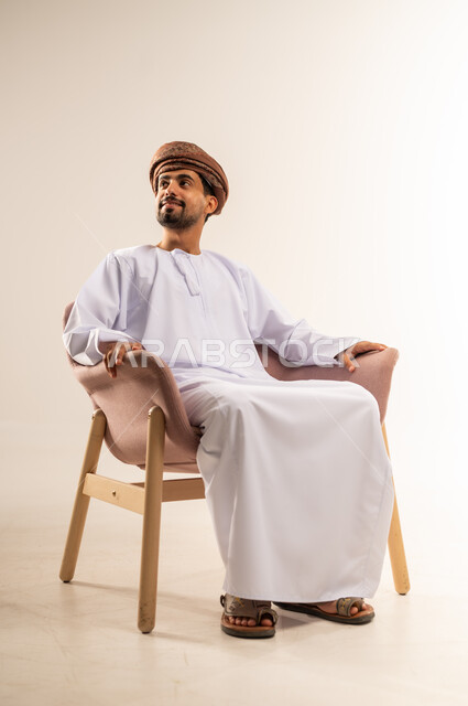 الاهتمام بالأناقة والمظهر الخارجي، مفهوم القوة والرجولة والشياكة، بورتريه لرجل عربي خليجي عماني مبتسم يرتدي الدشداشه والعمامة يجلس على كرسي خشبي مريح ينظر الى مكان ما في الاعلى بتعابير التفكير والتمعن والسرور ، ايماءات الثقة بالنفس، خلفية بيضاء