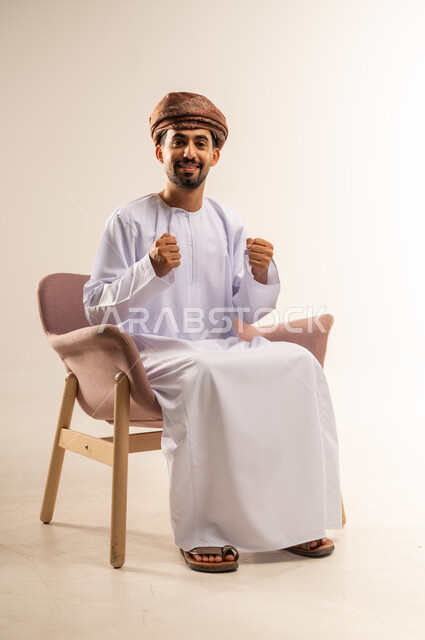 مفهوم الانجاز وتحقيق الاهداف، بورتريه لرجل عربي خليجي عماني مبتسم يرتدي الدشداشه والعمامة يجلس على كرسي مريح يرفع قبضة يديه للاعلى بإيماءات الفوز والانتصار والنجاح, النظر الى الكاميرا بتعابير السعادة والقوة والسرور، خلفية بيضاء