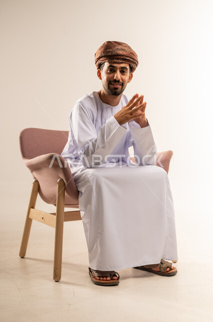 الاهتمام بالأناقة والمظهر الخارجي، مفهوم القوة والرجولة والشياكة، بورتريه لرجل عربي خليجي عماني مبتسم يرتدي الدشداشه والعمامة يجلس على كرسي خشبي مريح ينظر الى الكاميرا بتعابير الفرح والسرور ، ايماءات الثقة بالنفس، خلفية بيضاء