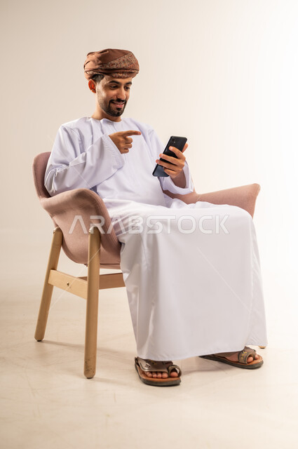 دمج التكنولوجيا في الحياة اليومية، استخدام التقنيات والأجهزة الحديثة، تصفح التطبيقات ومواقع التواصل الاجتماعي عبر الانترنت، دردشة ومحادثات وتعارف، بورتريه لرجل عربي خليجي عماني مبتسم يرتدي الدشداشة والعمامة يجلس على كرسي خشبي مريح يقوم باستخدام الهاتف المحمول، خلفية بيضاء