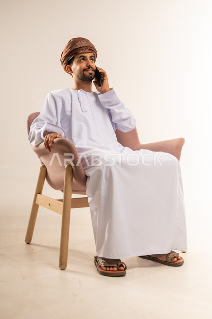 Benefiting from mobile services and applications, integrating technology and modern devices into daily life, portrait of an Arab Gulf Omani man wearing a dishdasha and turban sitting on a comfortable chair making a phone call using a mobile phone, communicating with family and friends, chatting, conversations and getting to know each other, white background