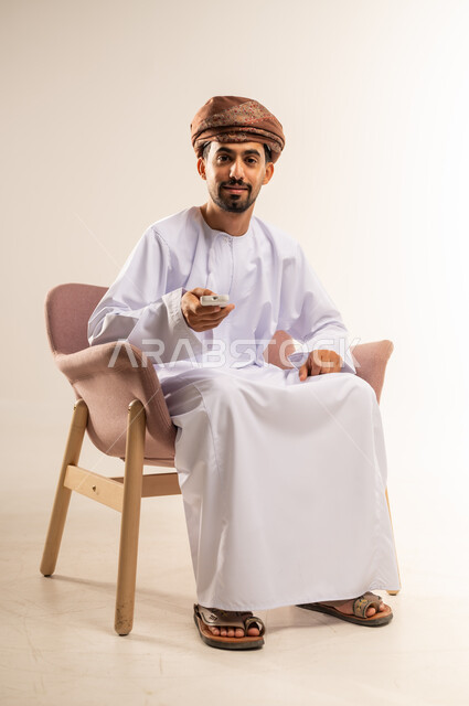 التغيير والتقليب بين القنوات، مفهوم التوجيه والتحكم، استخدام الاجهزة التقية الحديثة، بورتريه لرجل عربي خليجي عماني يرتدي الدشداشة والعمامة يجلس على كرسي مريح يحمل بيده جهاز التحكم، متابعة الافلام والبرامج المسلية، خلفية بيضاء