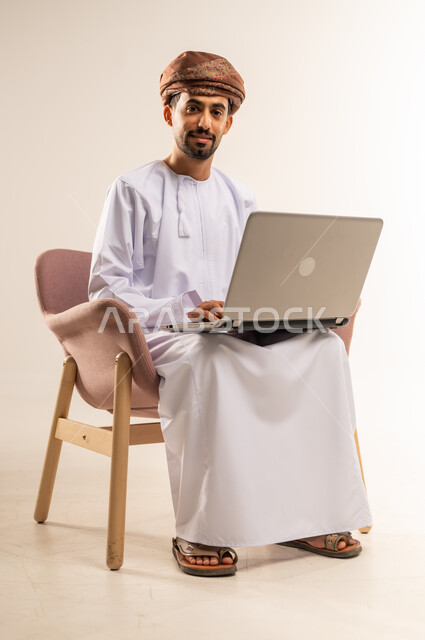 استخدام التقنيات الحديثة المتطورة، إدارة شؤون الأعمال وتنظيمها، بورتريه لرجل عربي خليجي عماني يرتدي الدشداشة والعمامة يجلس على كرسي مريح يعمل على جهاز الحاسوب المحمول, ايماءات السرور والسعادة, خلفية بيضاء