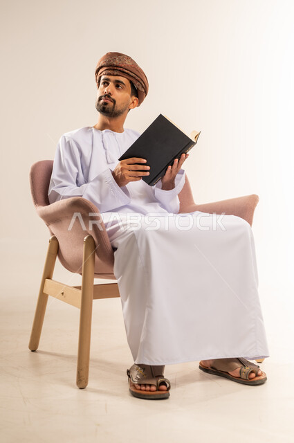 Love of reading and studying, spending free time learning and educating, portrait of an Arab Gulf Omani man wearing a dishdasha and turban sitting on a comfortable sofa holding a book in his hand, looking somewhere with gestures of concentration, interest in browsing useful books, white background