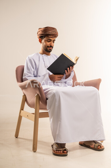 Spending free time learning and educating, love of reading, portrait of an Arab Gulf Omani man wearing a dishdasha and turban sitting on a comfortable sofa holding a book in his hand, interest in browsing useful books, white background