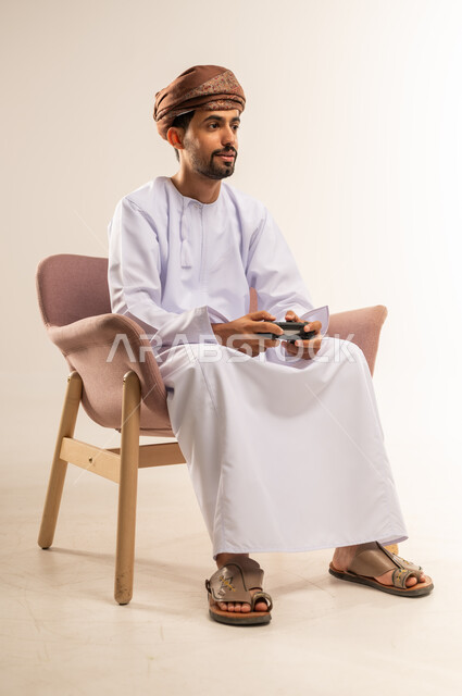 انشطة ترفيهية منزلية، قضاء وقت ممتع في المنزل، استخدام التكنولوجيا الحديثة في الترفيه، بورتريه لرجل عربي خليجي عماني يجلس على كرسي مريح يمسك بيده عصا التحكم يقوم بلعب العاب الفيديو بإيماءات التركيز، المتعة والتسلية والتفاعل، خلفية بيضاء