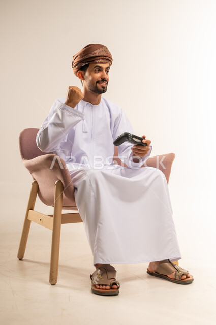 المتعة والتسلية والتفاعل، انشطة ترفيهية منزلية، قضاء وقت ممتع في المنزل، استخدام التكنولوجيا الحديثة في الترفيه، بورتريه لرجل عربي خليجي عماني يجلس على كرسي مريح يمسك بعصا التحكم يرفع يده بايماءات النصر، لعب العاب الفيديو بإيماءات السرور والسعادة، خلفية بيضاء