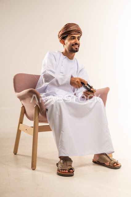 قضاء وقت ممتع في المنزل، انشطة ترفيهية منزلية، استخدام التكنولوجيا الحديثة في الترفيه، بورتريه لرجل عربي خليجي عماني يجلس على كرسي مريح يمسك بيده عصا التحكم يقوم بلعب العاب الفيديو بإيماءات السعادة والسرور، المتعة والتسلية والتفاعل، خلفية بيضاء