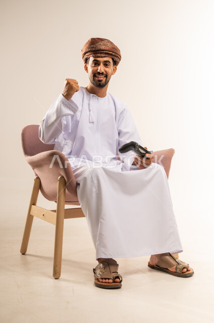 لعب العاب الفيديو بإيماءات السرور والسعادة، المتعة والتسلية والتفاعل، انشطة ترفيهية منزلية، قضاء وقت ممتع في المنزل، استخدام التكنولوجيا الحديثة في الترفيه، بورتريه لرجل عربي خليجي عماني يجلس على كرسي مريح يمسك بعصا التحكم يرفع يده بايماءات النصر، خلفية بيضاء