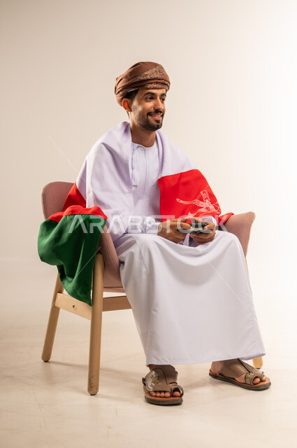 مفهوم الربح والفوز، الاحتفال بذكرى اليوم الوطني، التفاعل والاندماج مع ألعاب الفيديو، الفخر بالانتماء للبلاد، بورتريه لشاب عربي خليجي عماني يرتدي الدشداشة والعمامة يضع علم سلطنة عمان على ظهره يجلس على كرسي مريح يحمل عصا التحكم، خلفية بيضاء