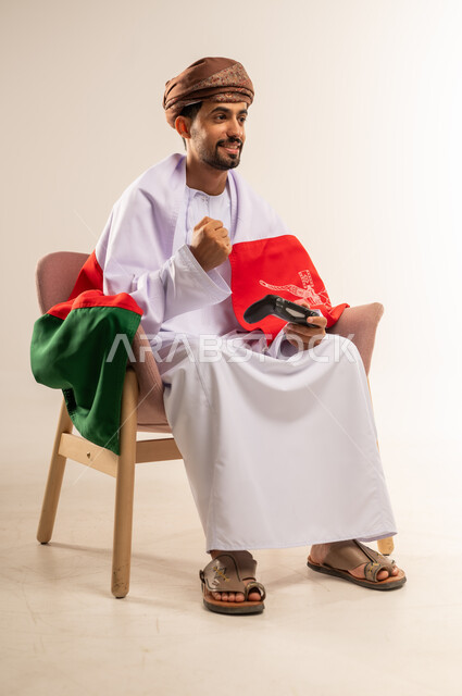 التفاعل والاندماج مع ألعاب الفيديو، الاحتفال بذكرى اليوم الوطني، الفخر بالانتماء للبلاد، بورتريه لشاب عربي خليجي عماني يرتدي الدشداشة والعمامة يضع علم سلطنة عمان على ظهره يجلس على كرسي مريح يحمل عصا التحكم يرفع يده بايماءات النصر، مفهوم الربح والفوز، خلفية بيضاء