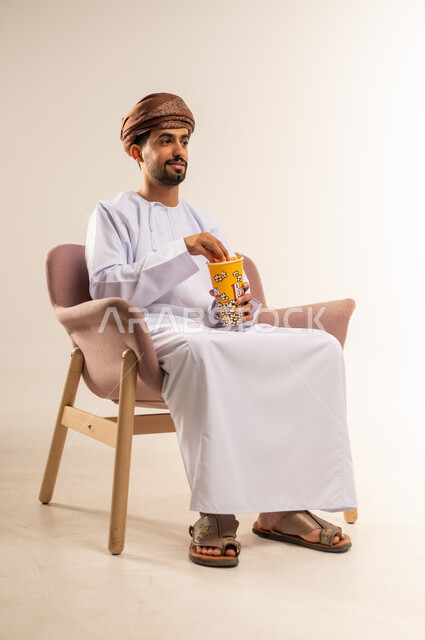 انشطة ترفيهية منزلية، الاستمتاع بتناول الفشار، بورتريه لرجل عربي خليجي عماني يرتدي الدشداشة والعمامة يجلس على كرسي مريح يحمل بيده علبة من الذرة يقوم بمتابعة التلفازبايماءات التركيز، سناكات ومقرمشات لذيذة، خلفيه بيضاء