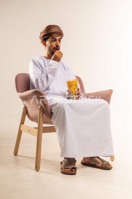 سناكات ومقرمشات لذيذة، انشطة ترفيهية منزلية، الاستمتاع بتناول الفشار، بورتريه لرجل عربي خليجي عماني يرتدي الدشداشة والعمامة يجلس على كرسي مريح يحمل بيده علبة من الذرة يقوم بمتابعة التلفازبايماءات الصدمة، خلفيه بيضاء