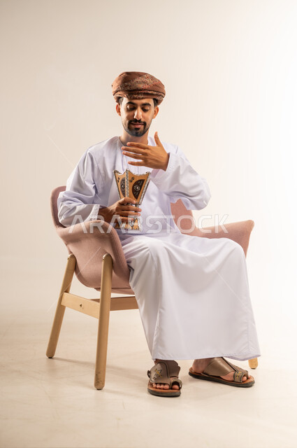 مفهوم التطيب بالبخور, استخدام خشب العود بالأعياد والمناسبات, العادات والتقاليد العمانية, بورتريه لرجل عربي خليجي عماني يرتدي الدشداشة والعمامة يجلس على كرسي مريح يقوم بالتعطر من المبخرة, الاهتمام بالمظهر الخارجي, خلفية بيضاء