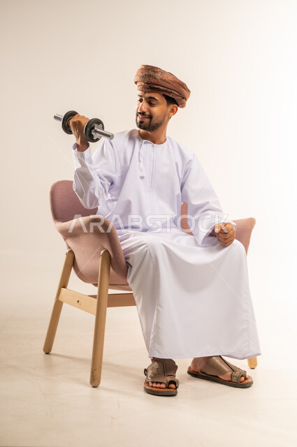 نشاط بدني للمحافظة على اللياقة الجسدية، رفع الأوزان الحديدية الثقيلة، بناء عضلات الجسم وتقويتها، بورتريه لرجل عربي خليجي عماني يرتدي الدشداشة والعمامة يجلس على كرسي مريح يحمل بيده الأثقال، خلفية بيضاء