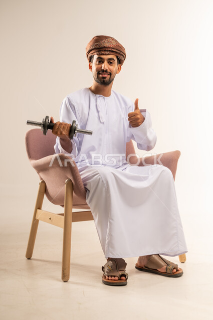 بناء عضلات الجسم وتقويتها، رفع الأوزان الحديدية الثقيلة، نشاط بدني للمحافظة على اللياقة الجسدية، بورتريه لرجل عربي خليجي عماني يرتدي الدشداشة والعمامة يجلس على كرسي مريح يحمل الأثقال يرفع اصبع الابهام بايماءات الاعجاب، خلفية بيضاء