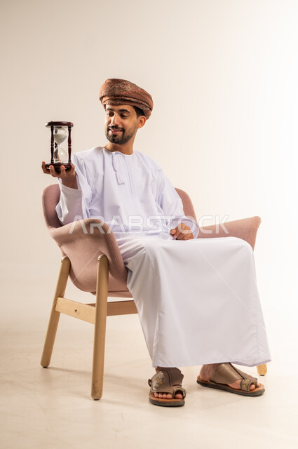 مفهوم إدارة الأعمال وتنظيم الجدول الزمني, ادراك قيمة الوقت، بورتريه لرجل عربي خليجي عماني يرتدي الدشداشة والعمامة يجلس على أريكة مريحة  يحمل ساعة رملية زجاجية (البنكام)، خلفية بيضاء
