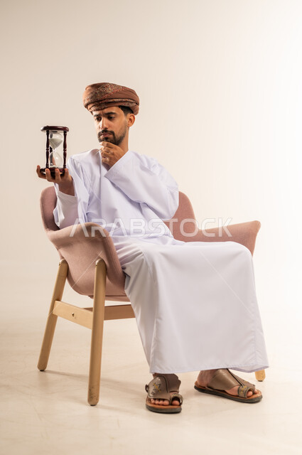 مفهوم إدارة الأعمال وتنظيم الجدول الزمني, ادراك قيمة الوقت، بورتريه لرجل عربي خليجي عماني يرتدي الدشداشة والعمامة يجلس على أريكة مريحة  يحمل ساعة رملية زجاجية (البنكام) يضع يده أسفل ذقنه بايماءات الشرود، خلفية بيضاء
