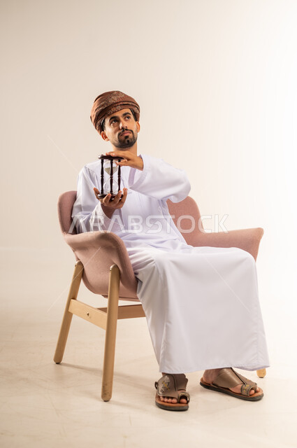 ادراك قيمة الوقت، مفهوم إدارة الأعمال وتنظيم الجدول الزمني, بورتريه لرجل عربي خليجي عماني يرتدي الدشداشة والعمامة يجلس على أريكة مريحة  يحمل ساعة رملية زجاجية (البنكام) ينظر الى مكانما بايماءات الشرود، خلفية بيضاء